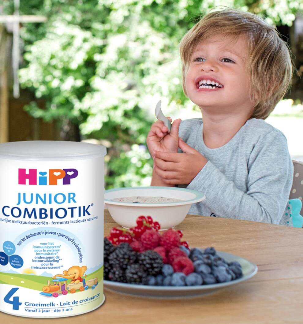 Een jong kind zit buiten aan tafel, met een lepel in de hand, met een potje Hipp Junior Combiotik poeder en een bord gemengde bessen op de voorgrond.