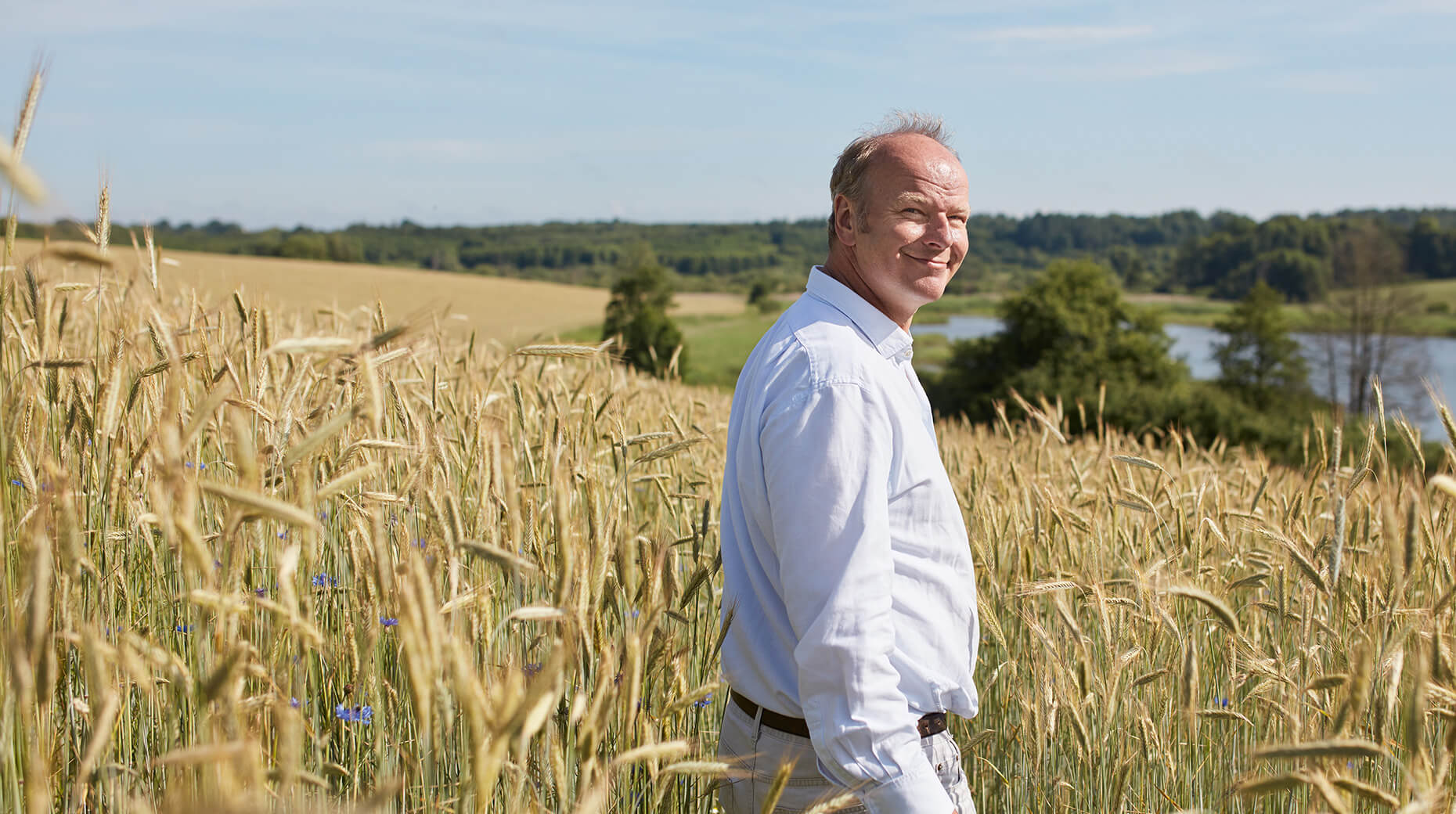 A man in a white shirt stands amidst a field of tall, golden wheat, with a scenic backdrop of a forested area and a water body.
