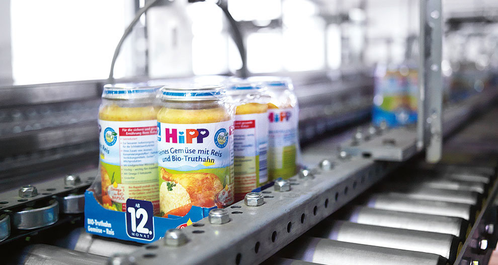 Een transportband in een fabriek transporteert potjes babyvoeding met het label "HiPP".