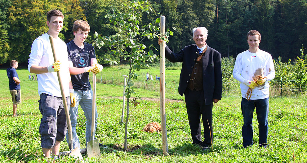 Vier personen staan in een grasveld met een jonge boom en tuingereedschap, wat duidt op een gemeenschappelijke tuinieractiviteit.