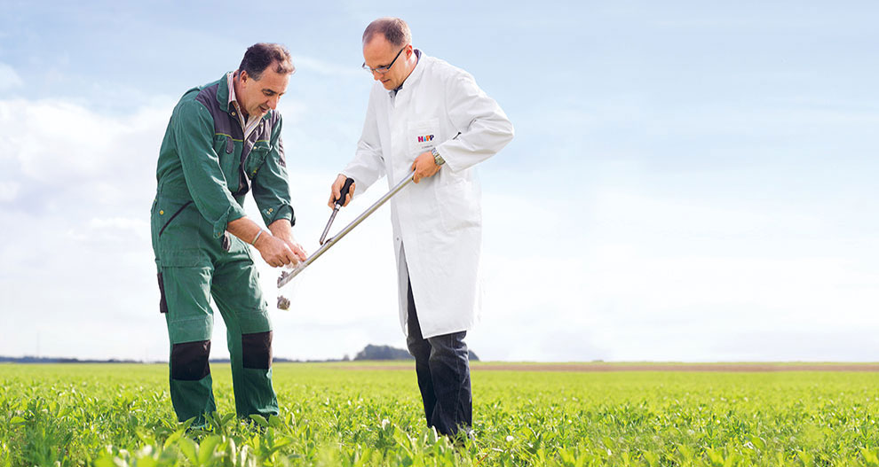 Twee mannen, de een in een witte laboratoriumjas en de ander in groene werkkleding, onderzoeken een bodemmonster in een veld.