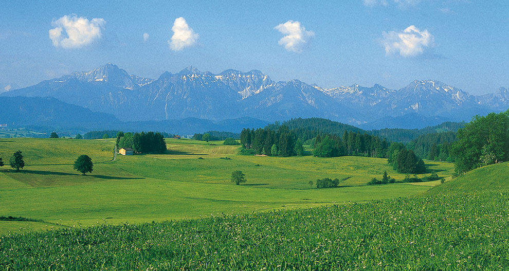 Een sereen landschap van glooiende groene velden, een eenzaam huis en majestueuze bergen onder een strakblauwe hemel.