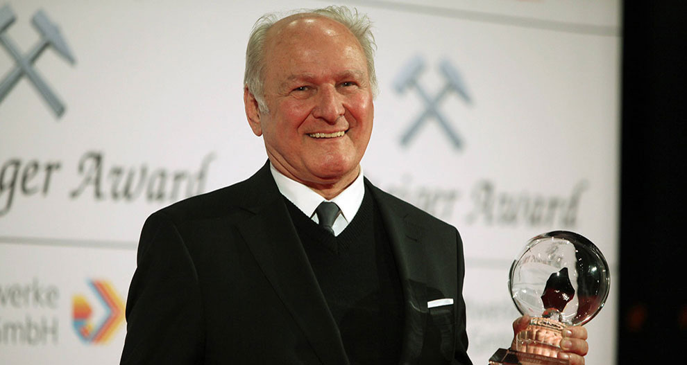Een oudere man in een zwart pak staat op een podium en houdt een doorzichtige bolvormige prijs vast, met de woorden "Schweizer Award" en logo's op de achtergrond.