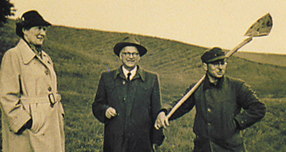 Drie personen staan in een open veld; één houdt een schep vast.