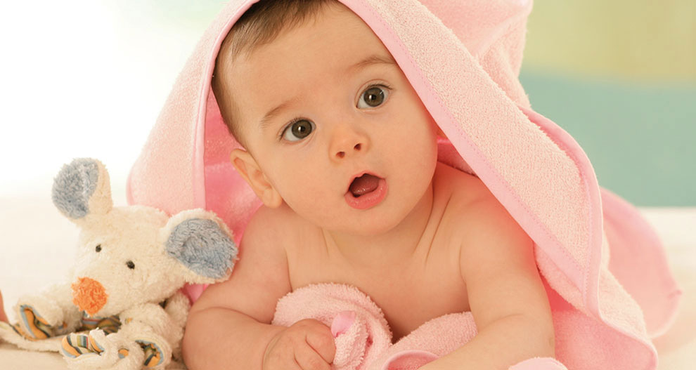 Een baby gewikkeld in een roze handdoek ligt op een wit oppervlak naast een pluchen speelgoedmuis.