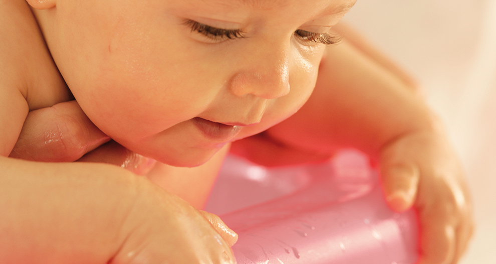 Een close-up van het gezicht en de handen van een baby, met waterdruppels op de huid, terwijl de baby in interactie is met een roze object.