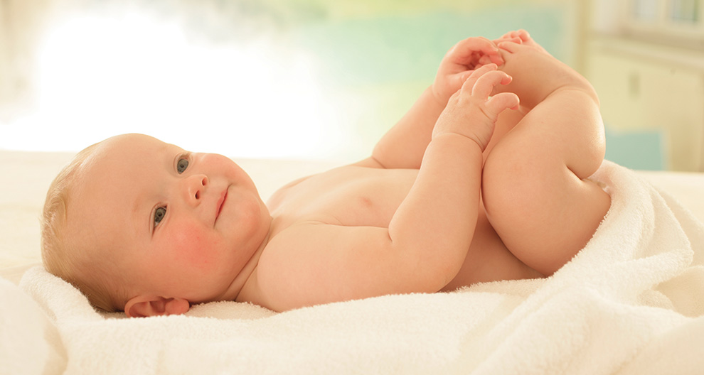 Een baby ligt op een witte deken en houdt zijn voetje vast.