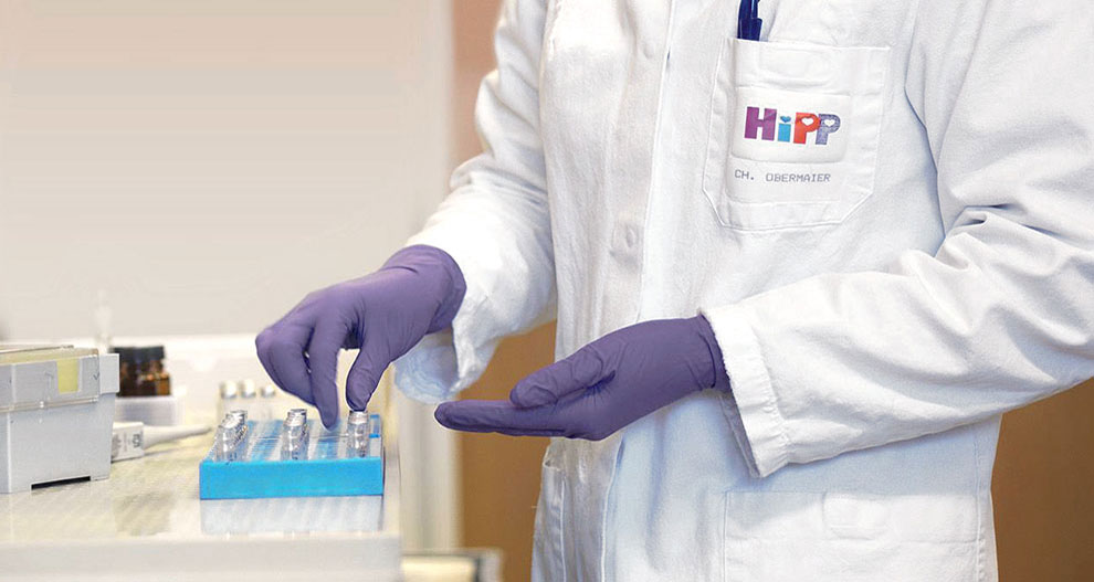 Een persoon in een witte laboratoriumjas met het logo "HiPP" en de naam "CH. OBERMAIER" draagt paarse handschoenen en hanteert een blauw dienblad met meerdere kleine containers.