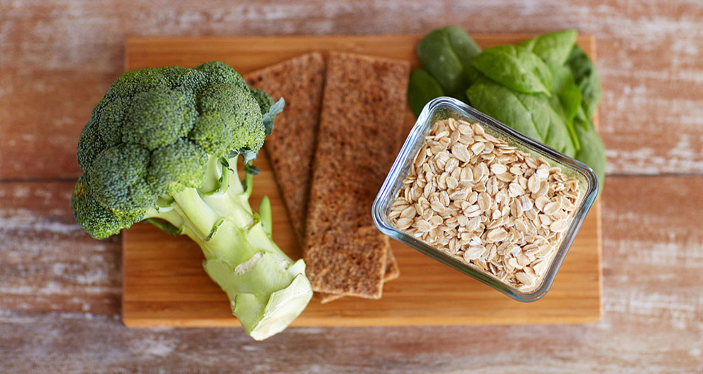 Op een houten snijplank liggen een stronk broccoli, een bos verse spinazie, een rechthoekig glazen bakje gevuld met havermout en twee rechthoekige bruine crackers.