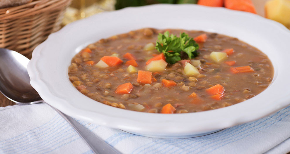 Een kom dikke, stevige soep met zichtbare stukken wortel, aardappel en linzen, gegarneerd met peterselie, vergezeld van een lepel en een mandje brood.