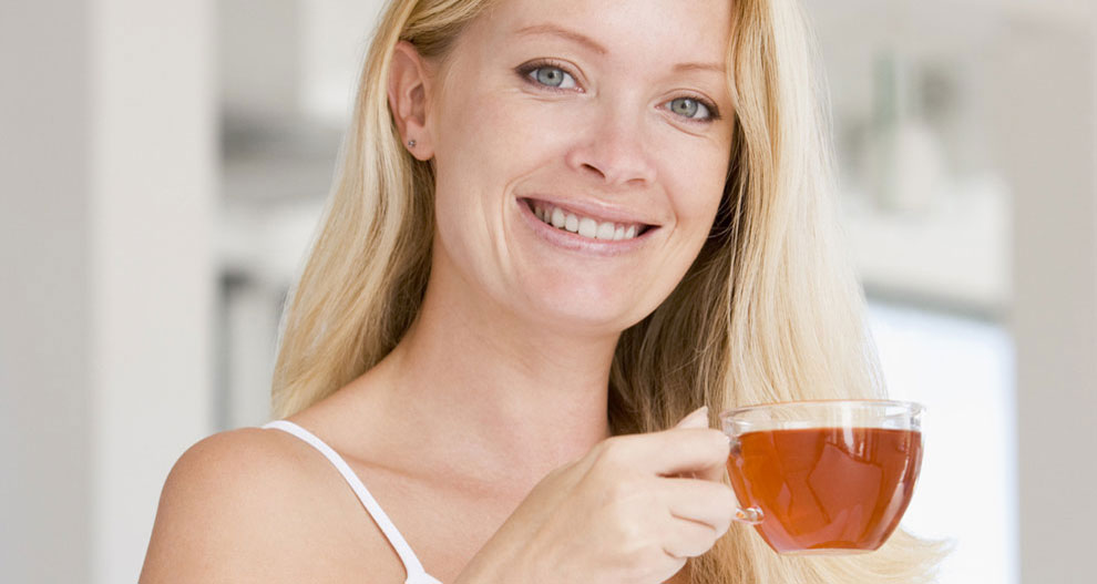 Een vrouw met blond haar houdt een helder glazen kopje vast gevuld met een bruine vloeistof, mogelijk thee of koffie.