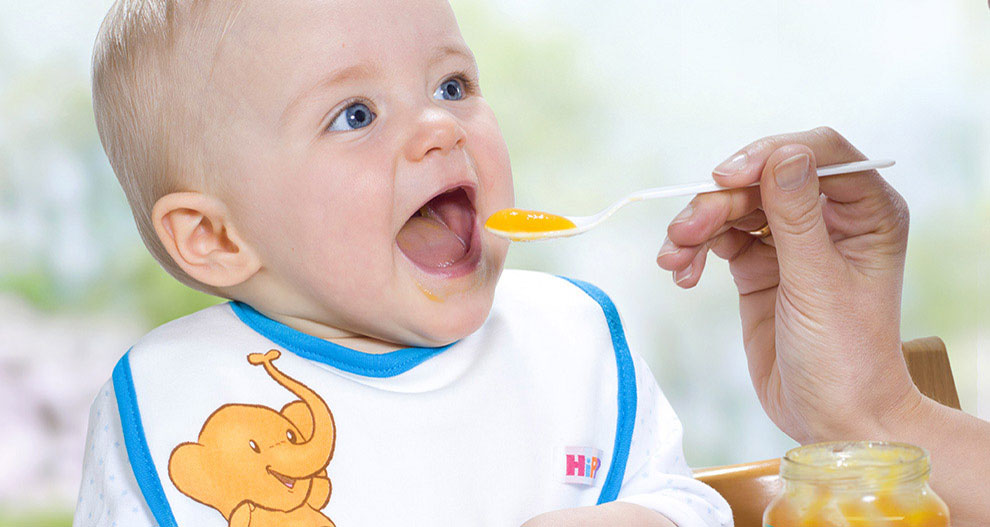 Een baby met een slabbetje wordt gevoed met een lepel uit een potje.