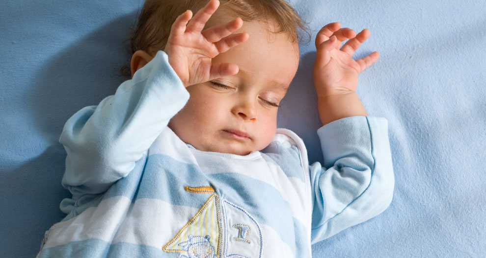 Een baby ligt op een blauwe ondergrond en draagt een lichtblauwe outfit met een zeilbootmotief.