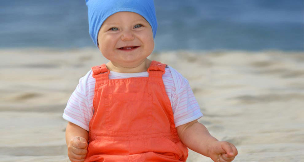 Een baby zit op een zandstrand en draagt een blauwe muts en een oranje overall.