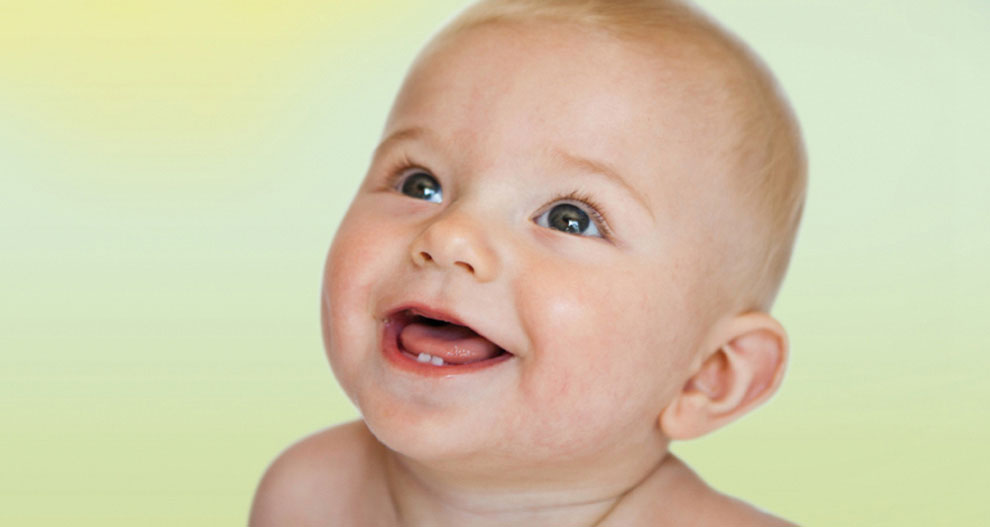 Een baby met een kaal hoofd, blauwe ogen en een lachende mond kijkt omhoog.