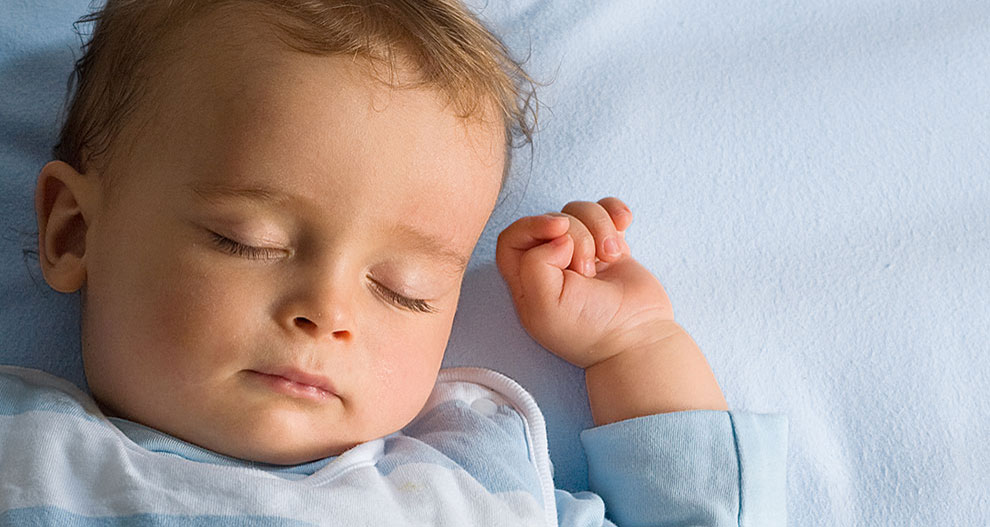 Een baby ligt op een blauw laken, draagt een lichtblauw en wit gestreept shirt, met één hand omhoog en gekrulde vingers.