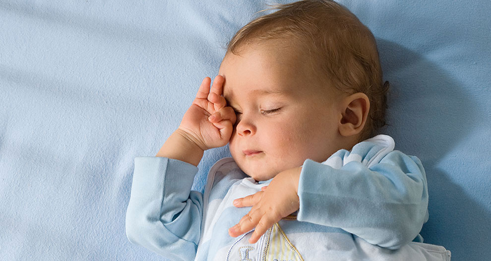 Een baby ligt op een blauw laken, draagt een lichtblauw pakje met een zeilbootmotief en bedekt één oog met een hand.