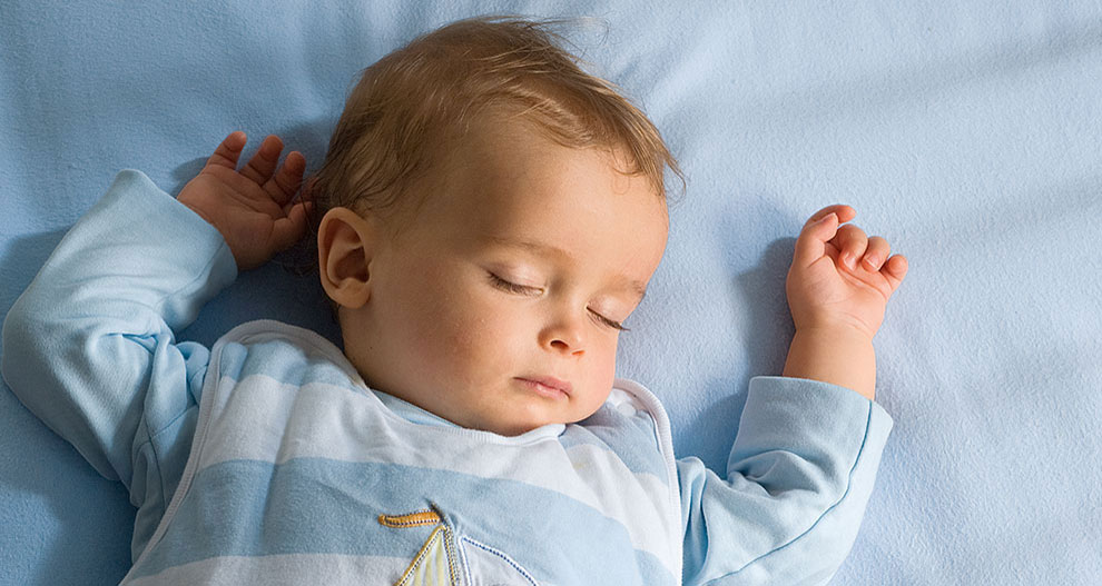 Een baby ligt op een blauw oppervlak en draagt een lichtblauw en wit gestreept pakje met een zeilbootmotief.