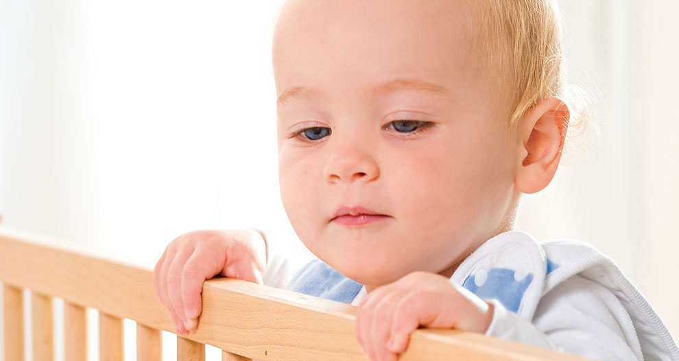 Een baby met licht haar en blauwe ogen tuurt over de reling van een houten wieg.