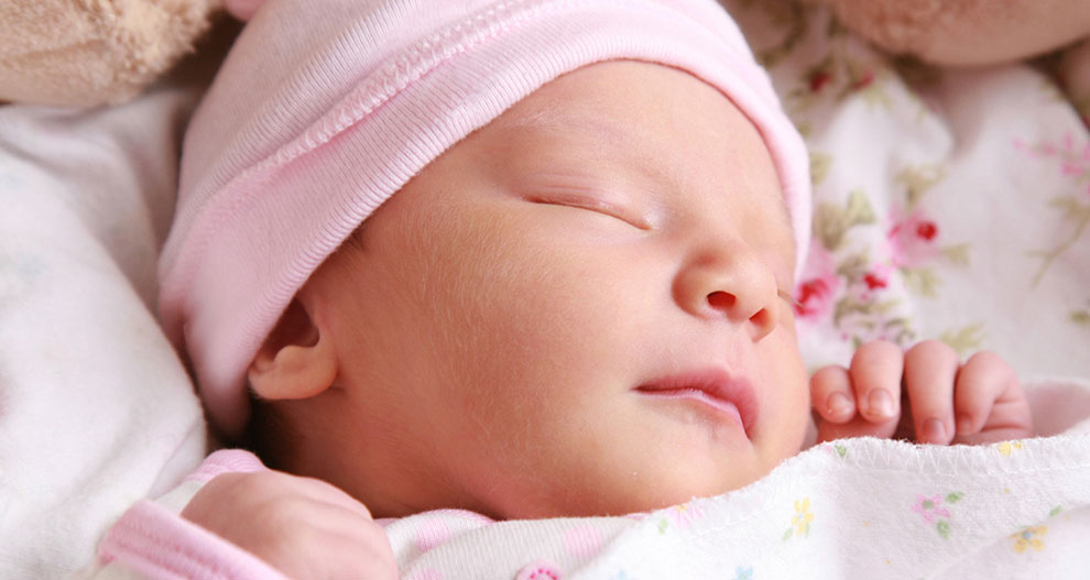 Een close-up van het gezicht van een slapende baby met een roze muts op en gewikkeld in een witte deken met bloemmotieven.