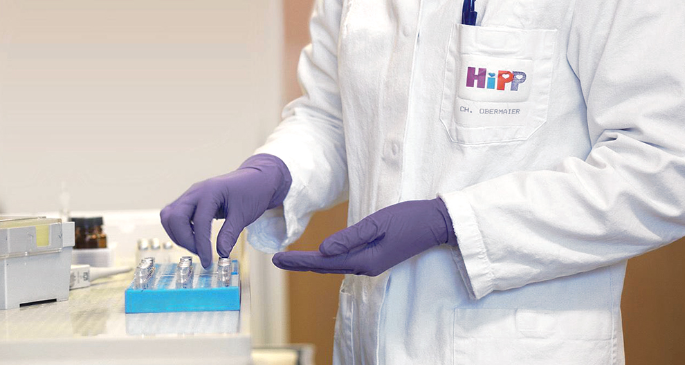 Een persoon in een witte laboratoriumjas met het logo "HiPP" en de naam "CH. OBERMAIER" hanteert een blauw dienblad met kleine flesjes.
