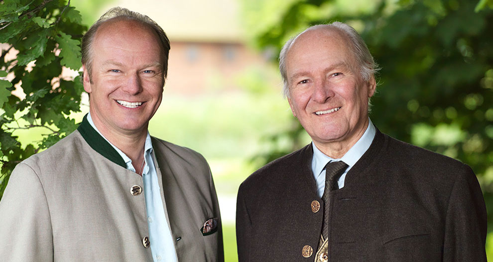 Twee mannen, de ene in een lichtgekleurde jas en de andere in een donkergekleurde jas, staan buiten naast elkaar.