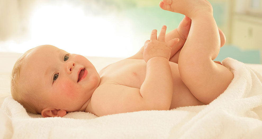 Een baby ligt op een witte handdoek en kijkt omhoog met een opgeheven hand.