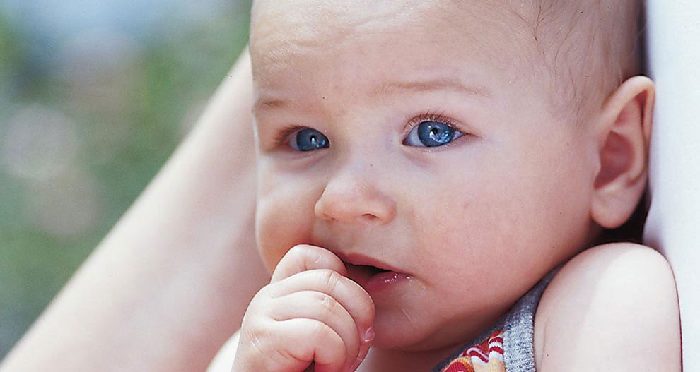 Een baby met blauwe ogen rust met zijn hoofd op iemands schouder en kauwt op zijn hand.