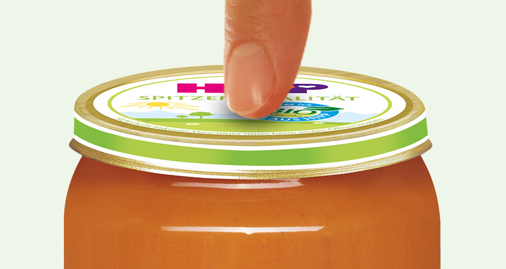 Een hand staat op het punt om een pot met oranjekleurige jam of conserven te openen met een groen en gouden deksel, waarop een etiket met een zon, bomen en het woord "BIO" staat.
