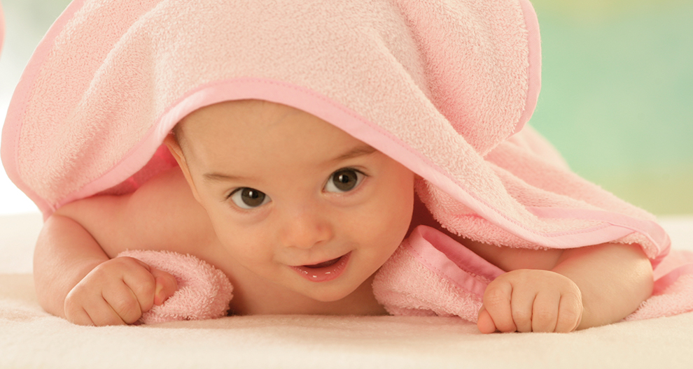 Een baby gewikkeld in een roze handdoek ligt op een wit oppervlak en kijkt recht in de camera.