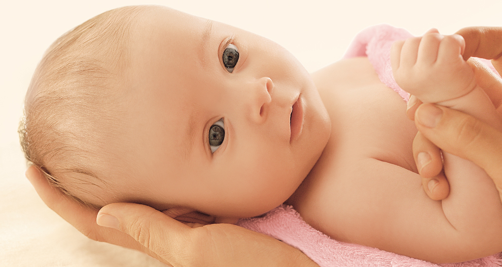 Een close-up van het gezicht en de handjes van een baby, zachtjes vastgehouden door de handen van een volwassene.