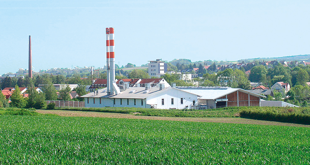 Een fabriek met een hoge schoorsteen staat in een groen veld en kijkt uit over een stad met huizen met rode daken en een hoge toren.