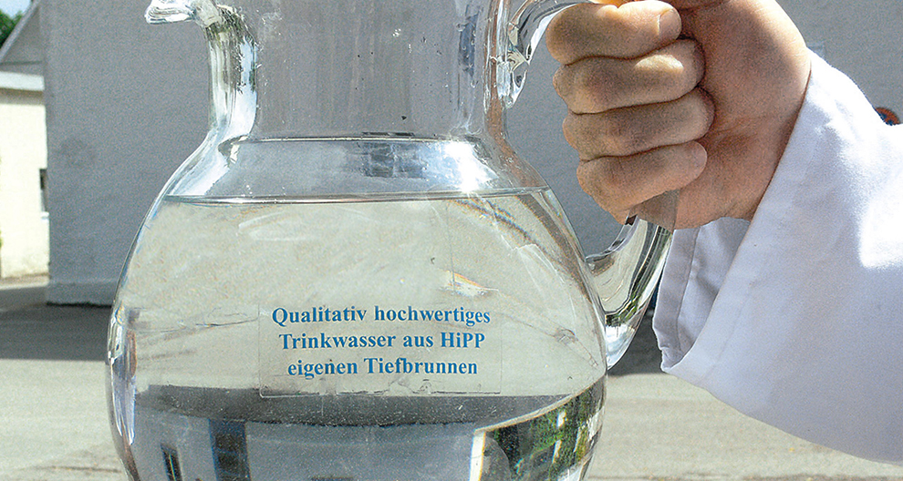 Een hand in een witte mouw houdt een helder glazen kan gevuld met water vast, met een label in het Duits op de kan.