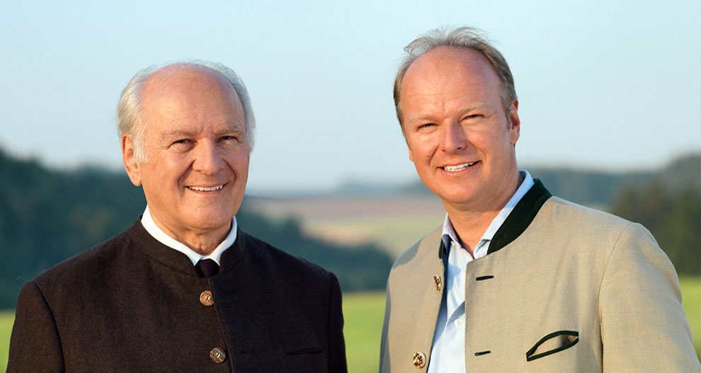 Twee mannen, een oudere met wit haar en een jongere met bruin haar, staan buiten naast elkaar.