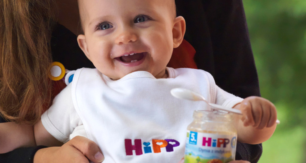Een baby met een wit slabbetje met het woord "HiPP" wordt gevoed uit een potje met een lepel.