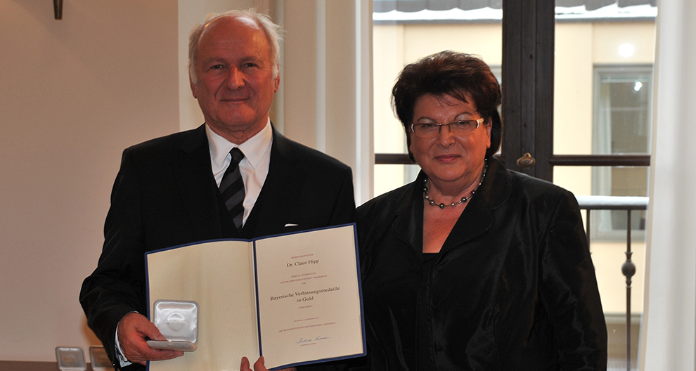 Een oudere man in een zwart pak houdt een certificaat en een rond zilveren voorwerp vast, staand naast een oudere vrouw in een zwarte outfit, beiden poseren binnenshuis met een raam op de achtergrond.