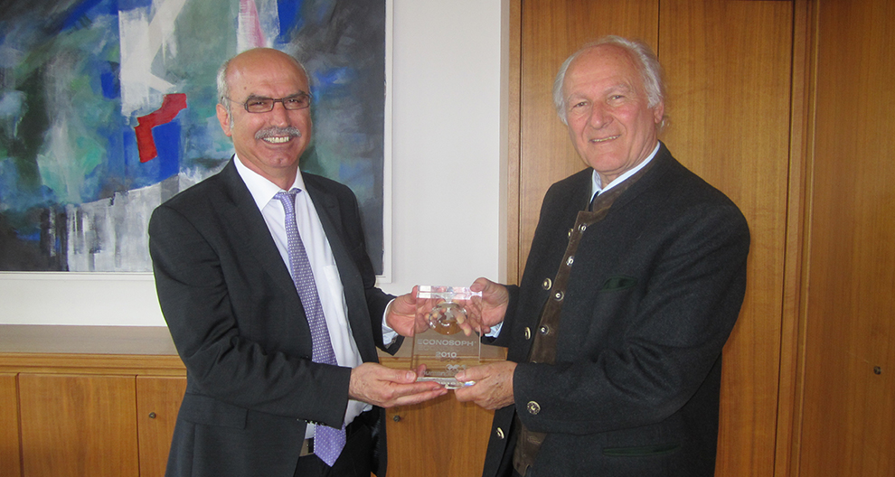 Twee mannen in pak staan in een kamer met een schilderij achter hen; de ene man overhandigt de andere een duidelijke prijs met het woord "ECONOSOPH" en het jaartal "2010" erop gedrukt.