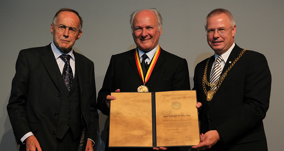 Drie mannen in formele kleding staan bij elkaar, waarbij de middelste figuur een groot, sierlijk certificaat en een medaille vasthoudt.