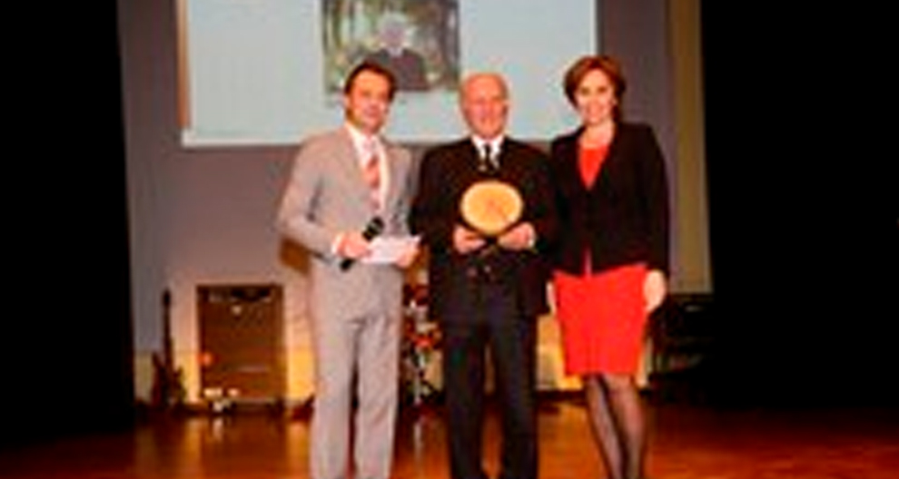 Drie personen staan op een podium, waarvan er één een ronde, houten prijs vasthoudt.