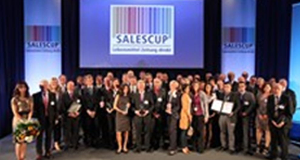 Een groep personen staat op een podium met een groot scherm waarop het woord "SALESCUP" en een afbeelding van een streepjescode worden weergegeven.