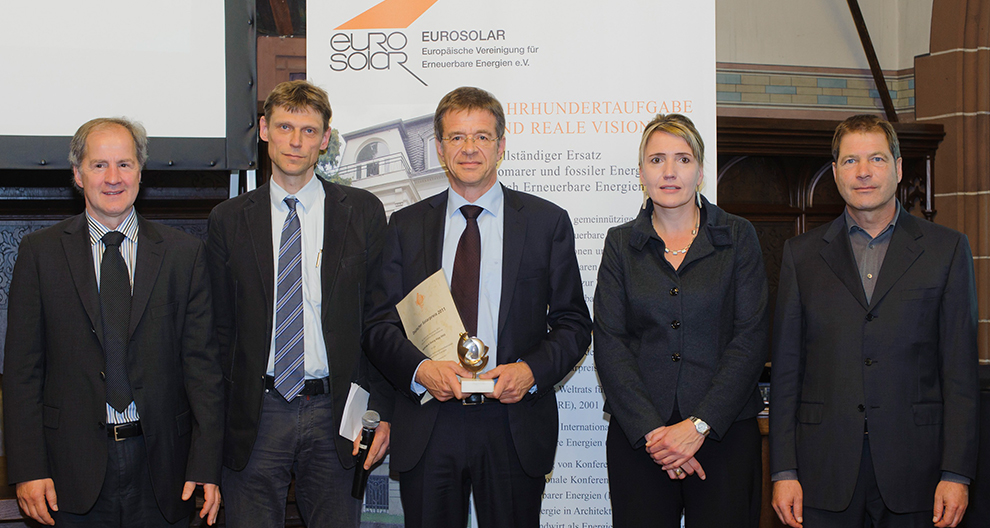 Vijf personen staan voor een reclamebanner voor 'Eurosolar' tijdens een evenement, waarbij één persoon een trofee en een certificaat vasthoudt.